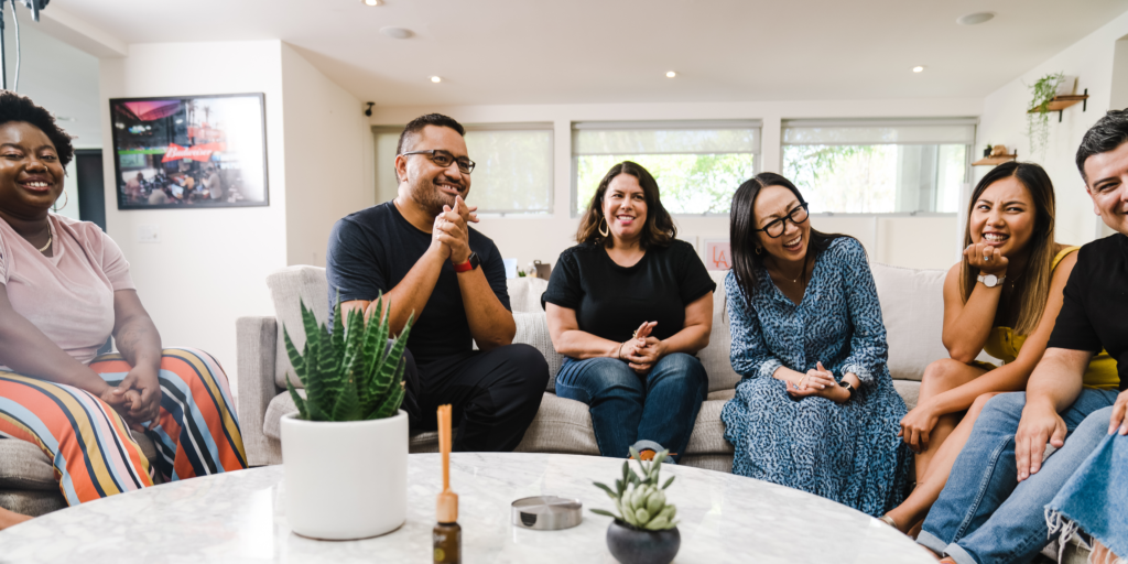 A group of 6 people sit on sofas around a table, they are chatting and smiling.