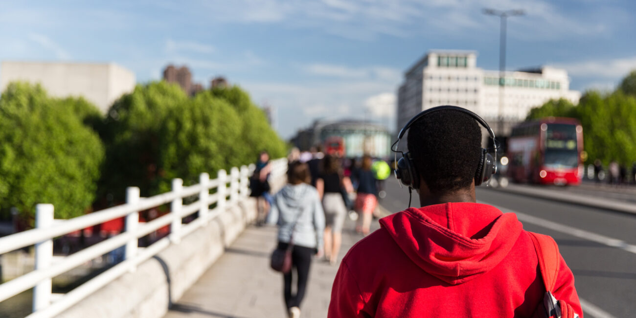 A young person wearing a red hoodie and headphones walks away from the camera on a busy Waterloo Bridge in London.